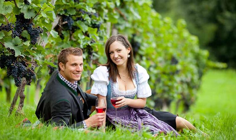 kostenloses KennenlernShooting: Paar im Weinberg, auf dem Boden sitzend mit vollen Rotweingläsern in den Händen