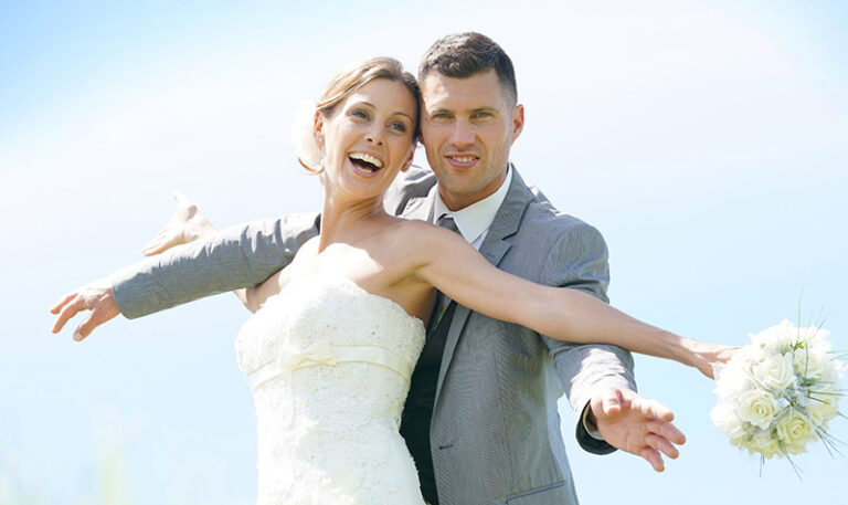 Strahlendlachendes Brautpaar die Arme seitlich von sich spreizend im Gegenlicht